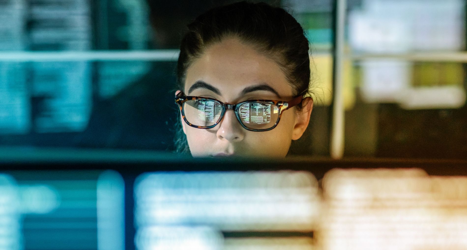 Frau mit Brille schaut in einem dunklen Raum in einen Bildschirm, vor und hinter ihr sieht man verschwommene Bildschirme.